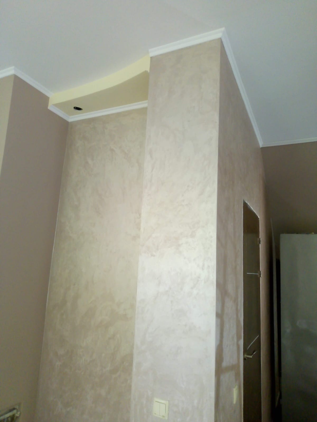 Wykończone wnętrze z dekoracyjną, beżową ścianą o nieregularnej fakturze, białe listwy przysufitowe, fragment drzwi i lodówki w tle.