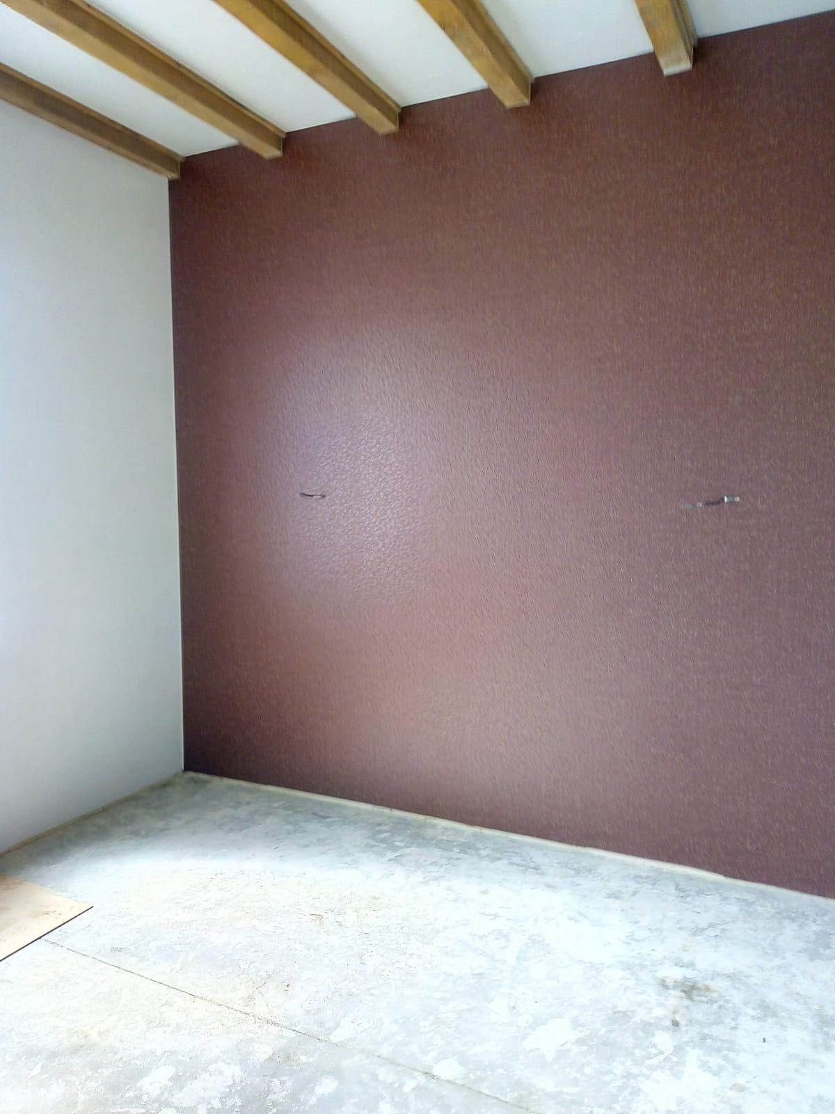 Pokój po remoncie: ściana w odcieniach brązu z delikatną fakturą, biała ściana boczna, surowa podłoga, drewniane belki sufitowe.