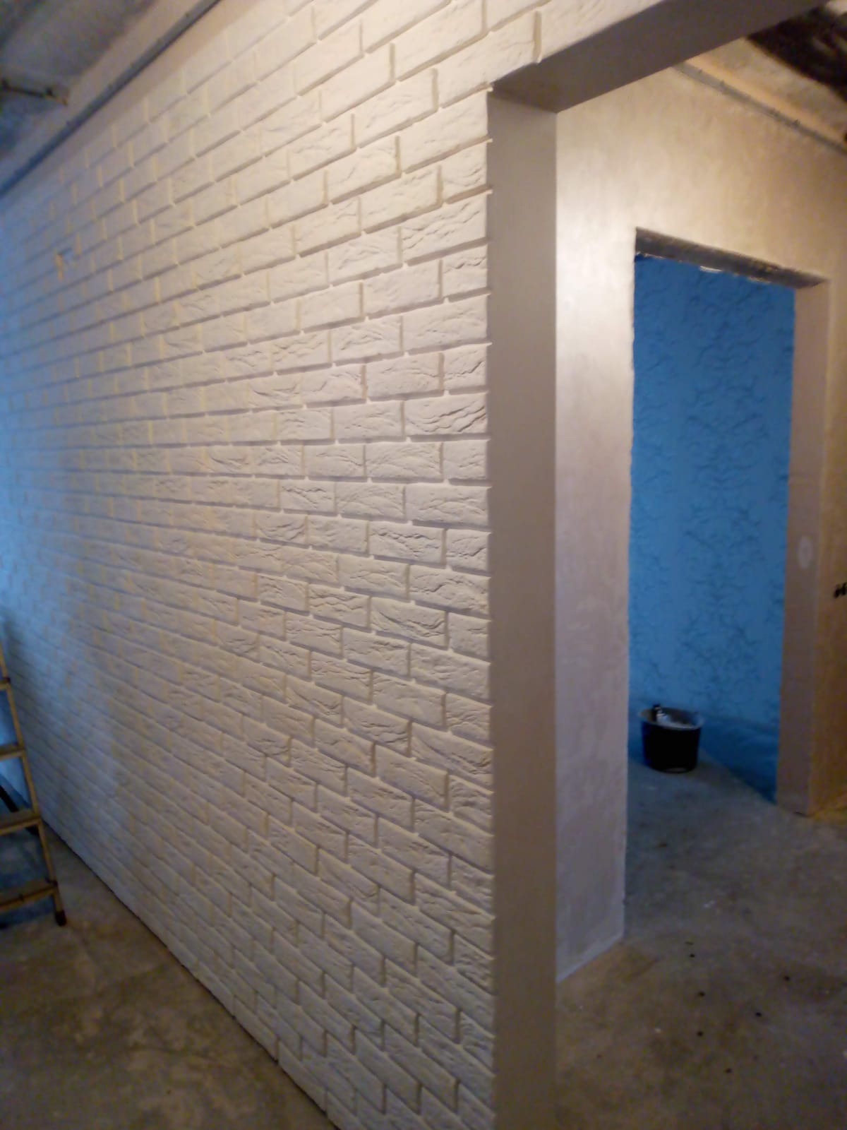 Ściana pokryta dekoracyjną, białą cegłą z widocznym wejściem do pomieszczenia z niebieską ścianą, ujęcie z boku.