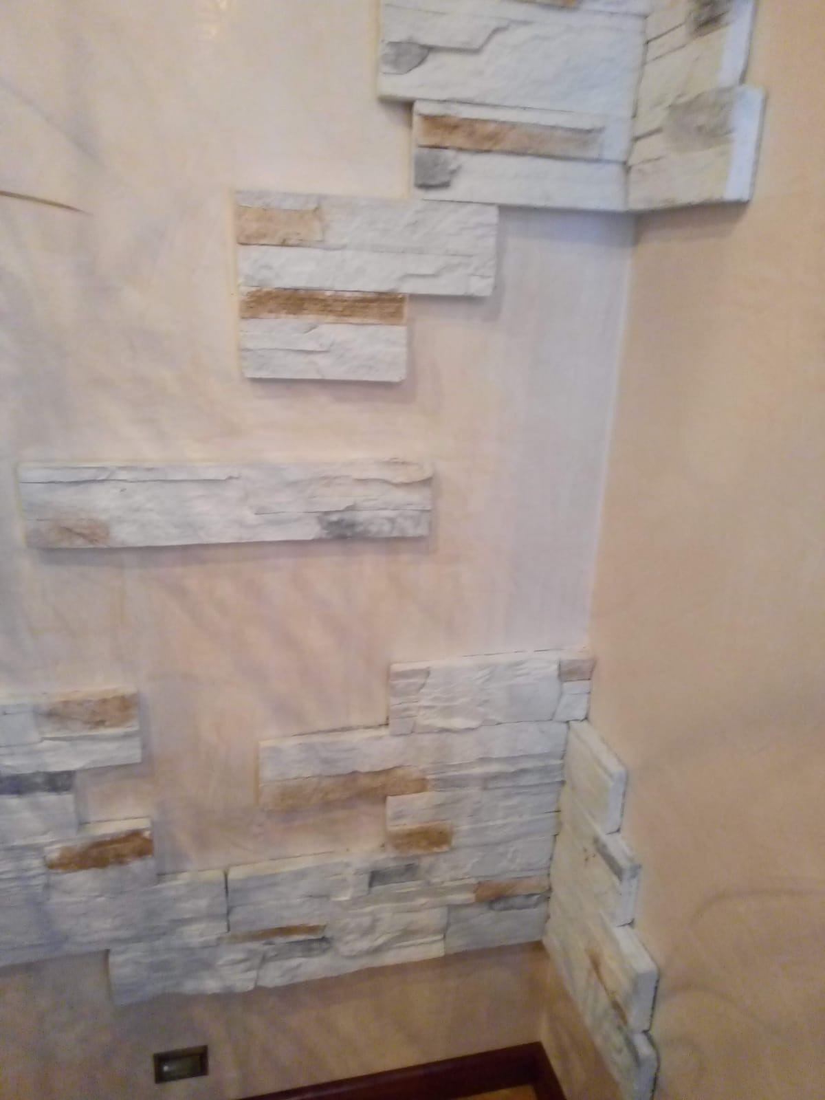 Fragment ściany wewnętrznej z dekoracyjnym wykończeniem z białych, nieregularnych płytek imitujących kamień, częściowo ułożonych na narożniku, na tle beżowej ściany z widoczną fakturą.