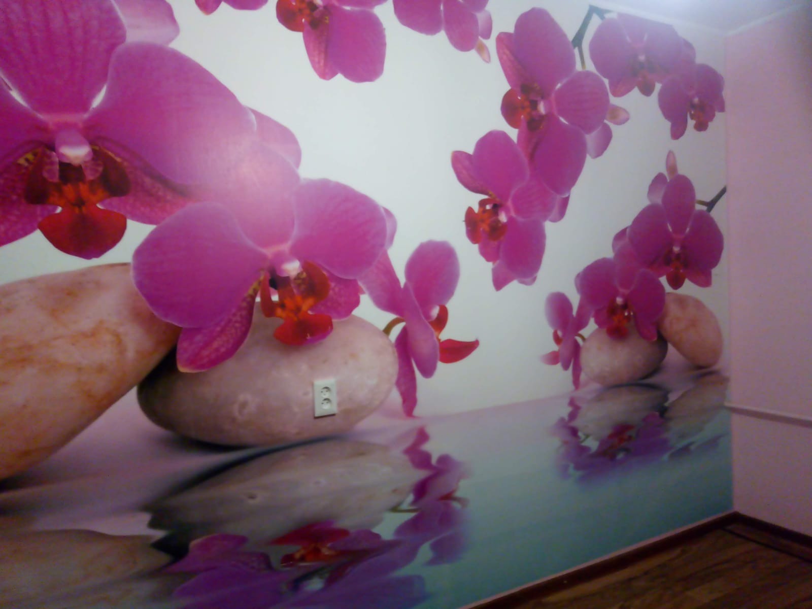 Fototapeta w rogu pokoju przedstawiająca różowe orchidee i kamienie z odbiciem w wodzie, z wkomponowanym gniazdkiem elektrycznym w jeden z kamieni.