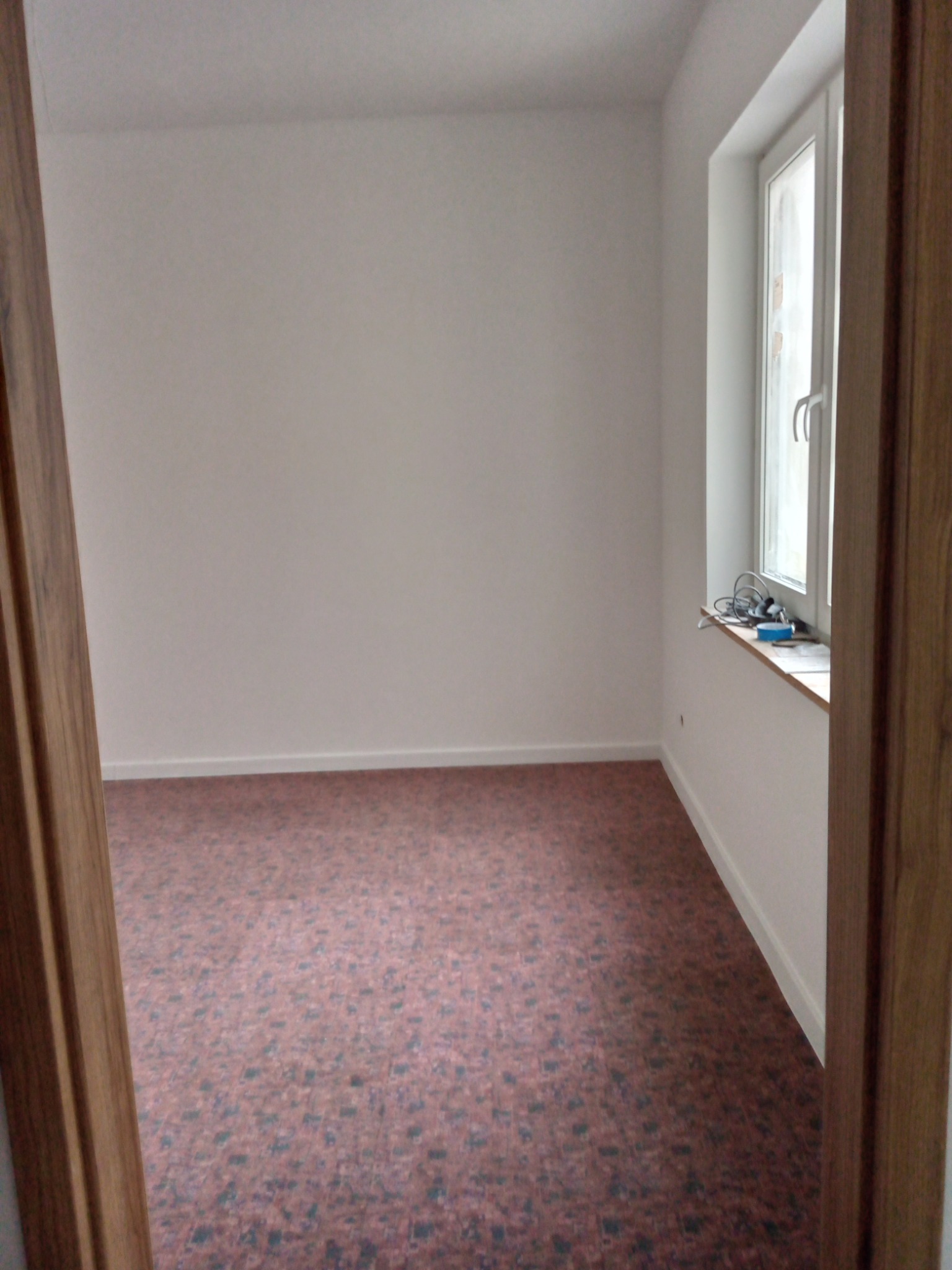 Pusty pokój z brązową wykładziną, świeżo pomalowanymi białymi ścianami i oknem z przewodami na parapecie, widok zza drewnianej futryny.