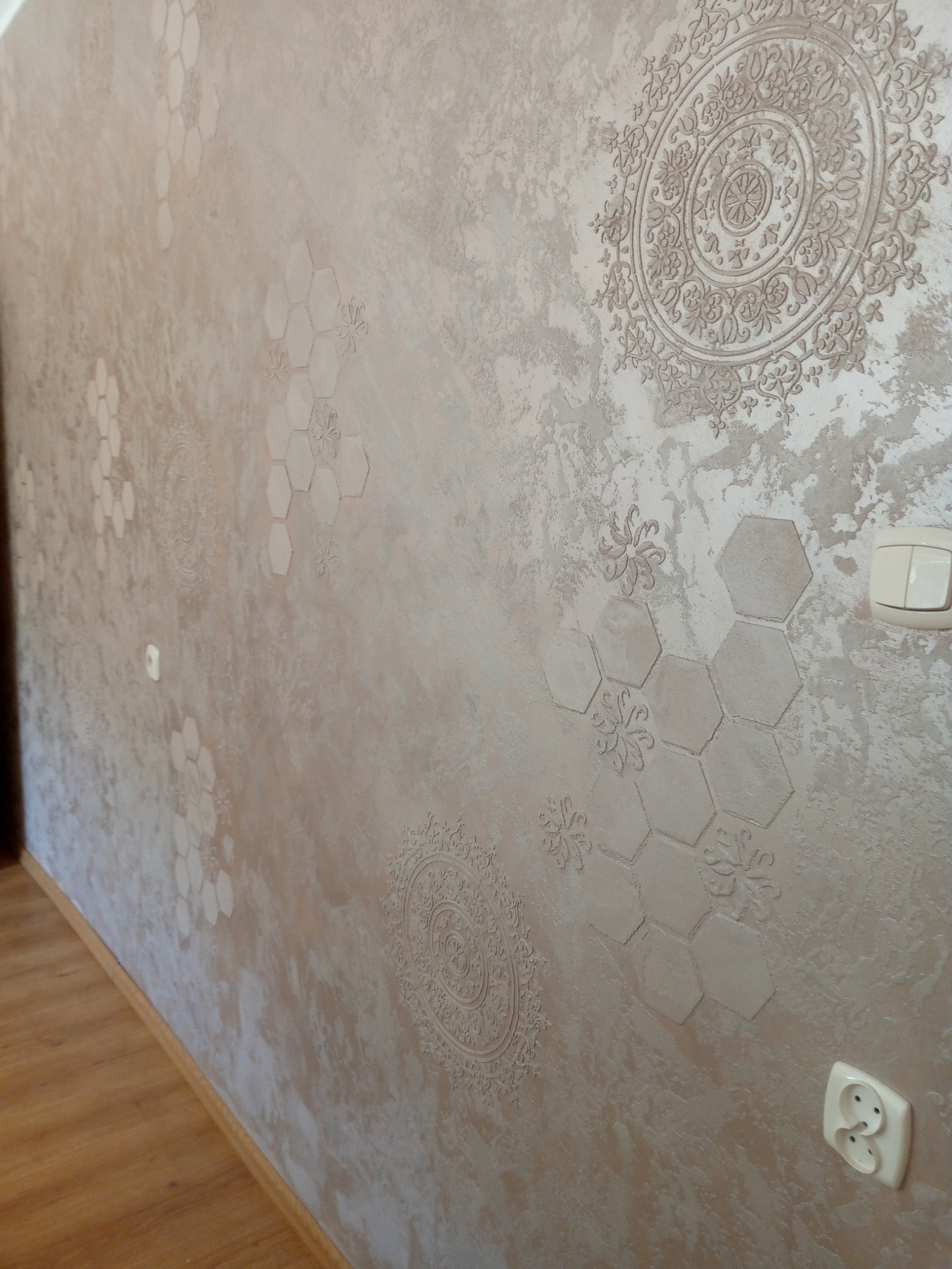 Dekoracyjna ściana w odcieniach beżu z wzorami mandali i heksagonów, wykonana techniką reliefową. Widoczne gniazdko elektryczne i fragment podłogi.