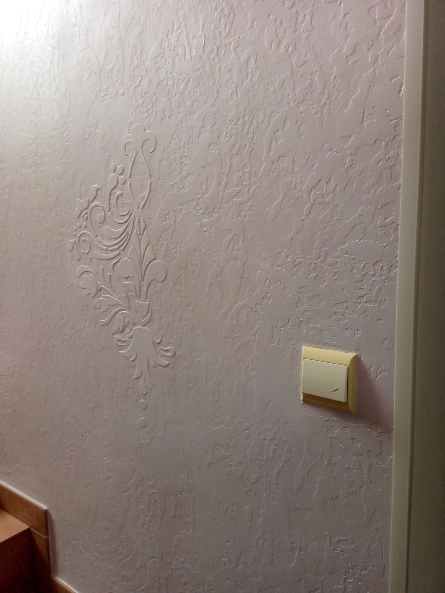 Ściana z dekoracyjną strukturą i wzorem ornamentu, oświetlona z góry. Obok włącznik światła i fragment futryny drzwi. Widoczne schody z brązowymi płytkami.