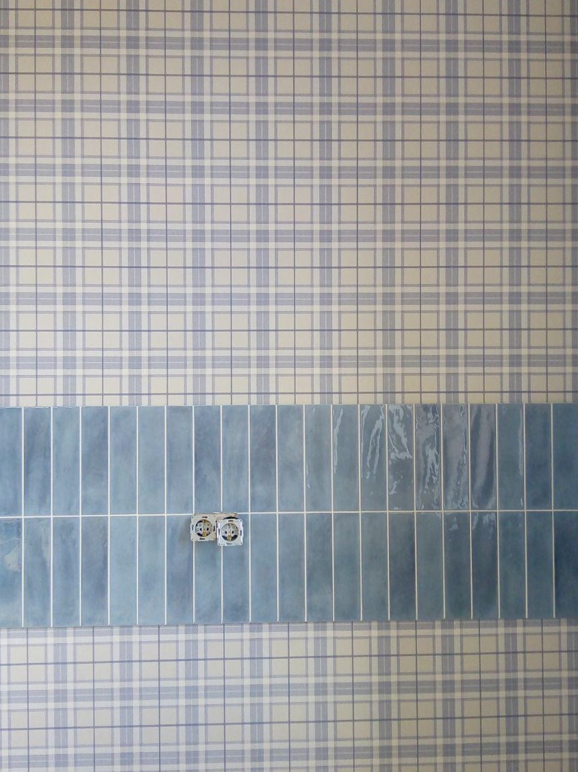 Ściana wykończona tapetą w kratę i pasem błękitnych, pionowych płytek, z widocznymi gniazdkami elektrycznymi. Jasne, naturalne oświetlenie.