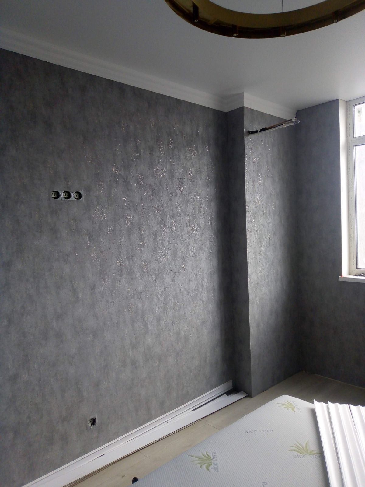 Szara ściana z dekoracyjną fakturą, listwy przypodłogowe w trakcie montażu, widoczne puszki elektryczne i fragment sufitu z oświetleniem.