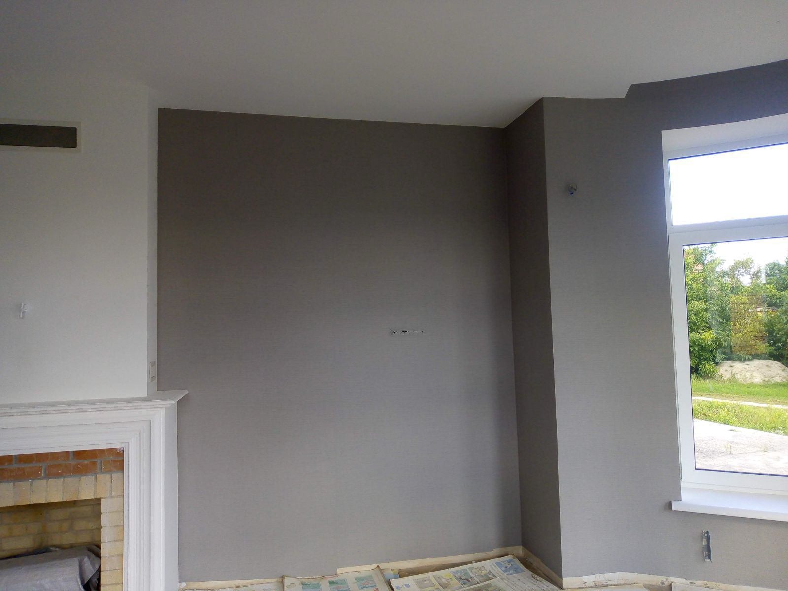 Świeżo pomalowane na szaro ściany pokoju z kominkiem, zabezpieczone podłogi gazetami. Widok na zewnątrz przez okno. Minimalistyczny design, spokojna kolorystyka.