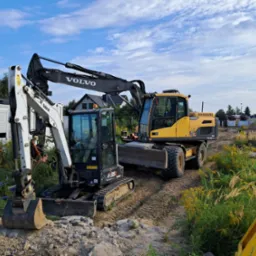 Dwie maszyny budowlane: biała mini koparka Bobcat E35z z przodu i żółta koparka kołowa Volvo EW160D z tyłu, stoją na placu budowy z widoczną stertą ziemi i trawami, pod błękitnym niebem.