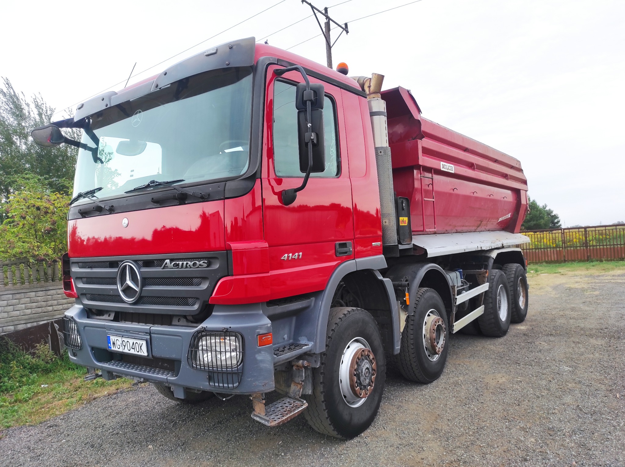 Czerwona wywrotka Mercedes Actros 4141 z widocznym numerem rejestracyjnym WG 9040K, zaparkowana na żwirowym placu, z zielenią w tle.