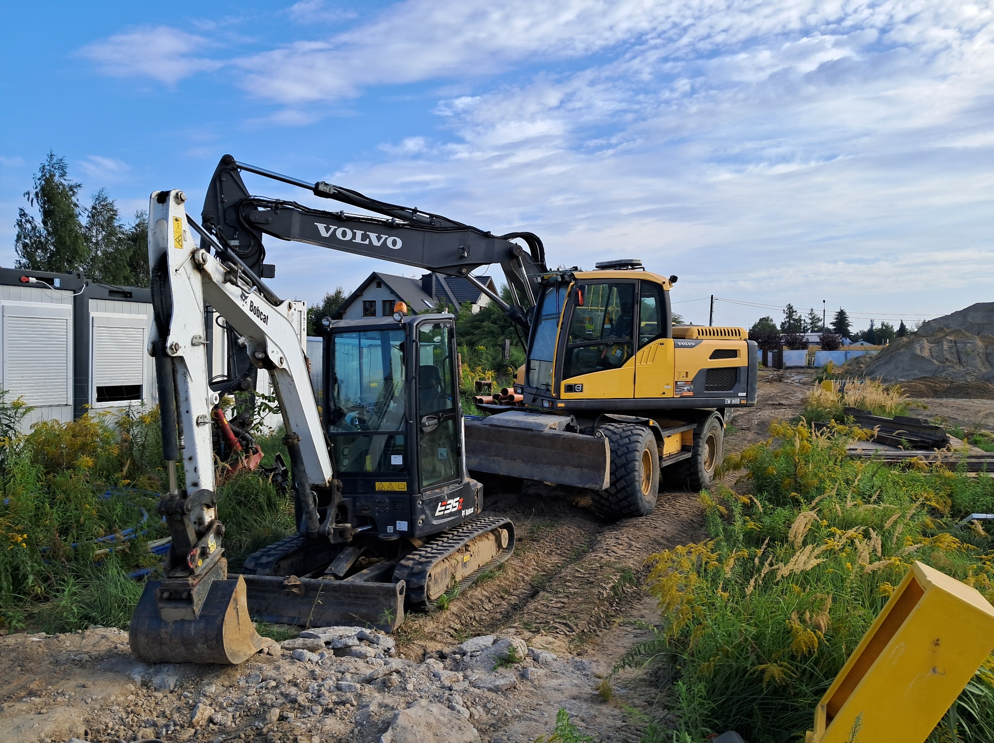 Dwie maszyny budowlane: biała mini koparka Bobcat E35z z przodu i żółta koparka kołowa Volvo EW160D z tyłu, stoją na placu budowy z widoczną stertą ziemi i trawami, pod błękitnym niebem.