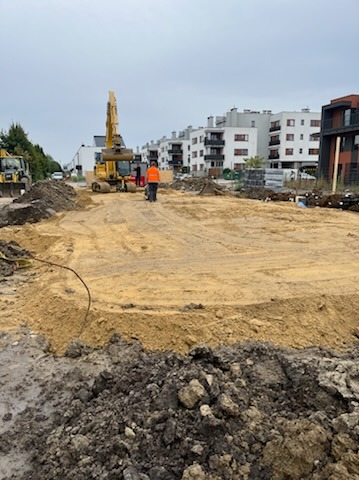 Żółta koparka gąsienicowa CAT na placu budowy, w tle budynki mieszkalne, widoczny pracownik w pomarańczowej kamizelce, świeżo wyrównany teren z widocznymi śladami opon.