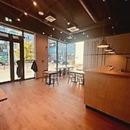 Remonty I wykonczenia - Jasne wnętrze biura lub lokalu usługowego z parkietem, dużymi oknami i widocznymi stolikami. Ściana w odcieniach szarości, czarny sufit z oświetleniem punktowym. Widoczny fragment lady recepcyjnej...