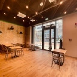 Remonty I wykonczenia - Jasne, przestronne wnętrze biura z drewnianą podłogą, stolikami i krzesłami, widok na ulicę przez duże okno, oświetlenie sufitowe punktowe, ściana w naturalnej tonacji z dekoracjami.