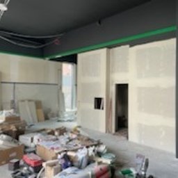 Remonty I wykonczenia - Wnętrze w trakcie prac wykończeniowych, widoczne ściany z płyt gipsowo-kartonowych z wyciętym otworem, sufit pomalowany na ciemny kolor, widoczne przewody, rusztowanie i materiały budowlane...