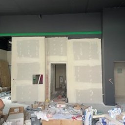 Remonty I wykonczenia - Montaż płyt kartonowo-gipsowych na ścianie z widocznymi spoinami i otworem na instalacje, w tle widoczny surowy stan pomieszczenia z rusztowaniem i materiałami budowlanymi.