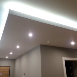 Remonty I wykonczenia - Sufit podwieszany z oświetleniem LED: listwa LED wzdłuż krawędzi i rząd punktowych lamp halogenowych, szara ściana z drzwiami w tle.