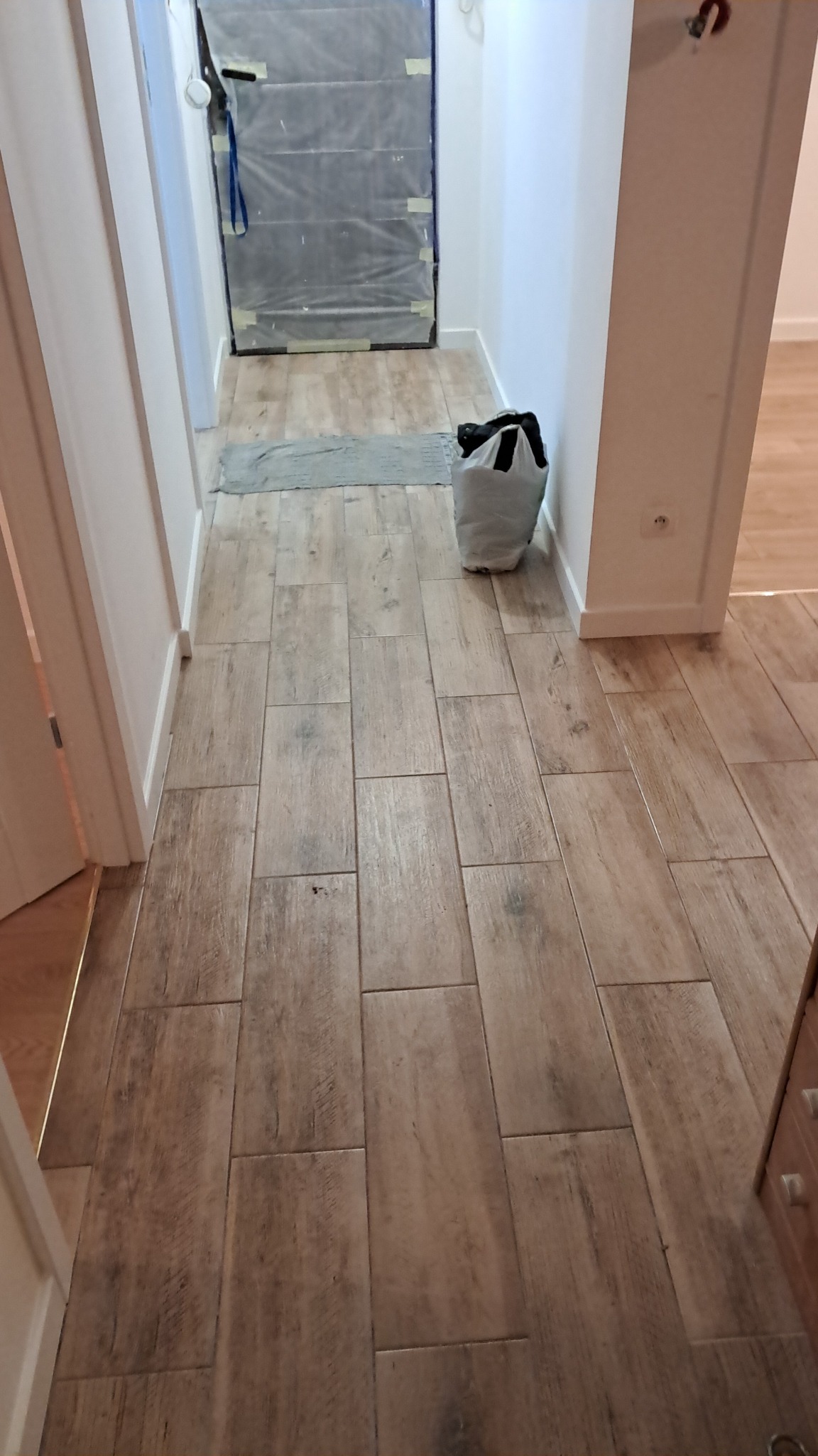 Korytarz w trakcie remontu z nową, drewnopodobną podłogą z płytek, zabezpieczone drzwi folią malarską i torba z narzędziami przy ścianie.