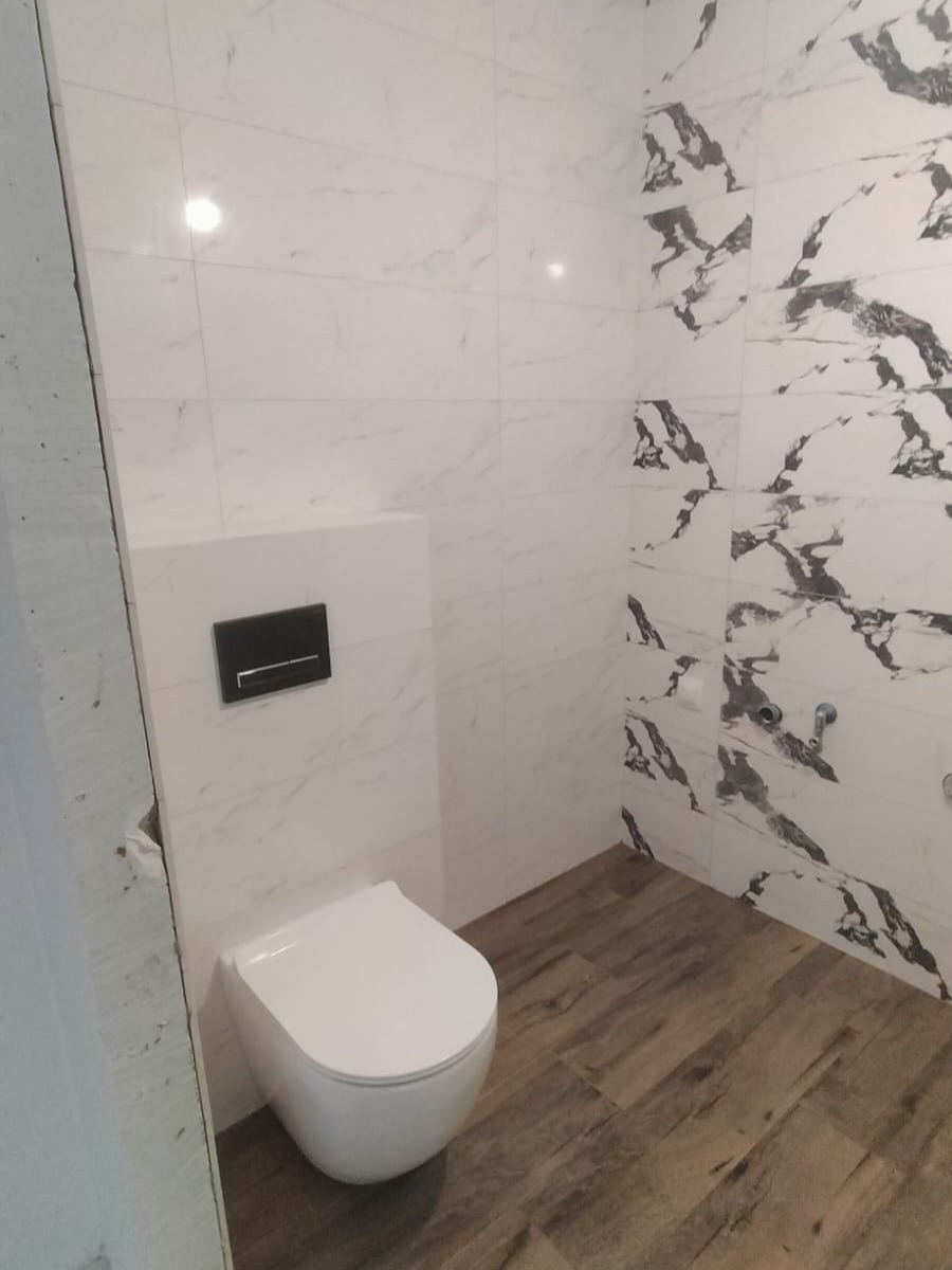 Nowoczesna toaleta podwieszana z czarnym przyciskiem spłukującym na ścianie wyłożonej białymi płytkami imitującymi marmur, uzupełniona podłogą z paneli imitujących drewno.