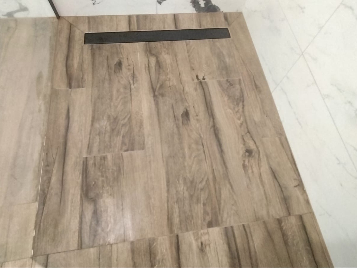 Podłoga w łazience wyłożona płytkami imitującymi drewno, z czarnym odpływem liniowym, sąsiadująca z białymi płytkami imitującymi marmur na ścianie.