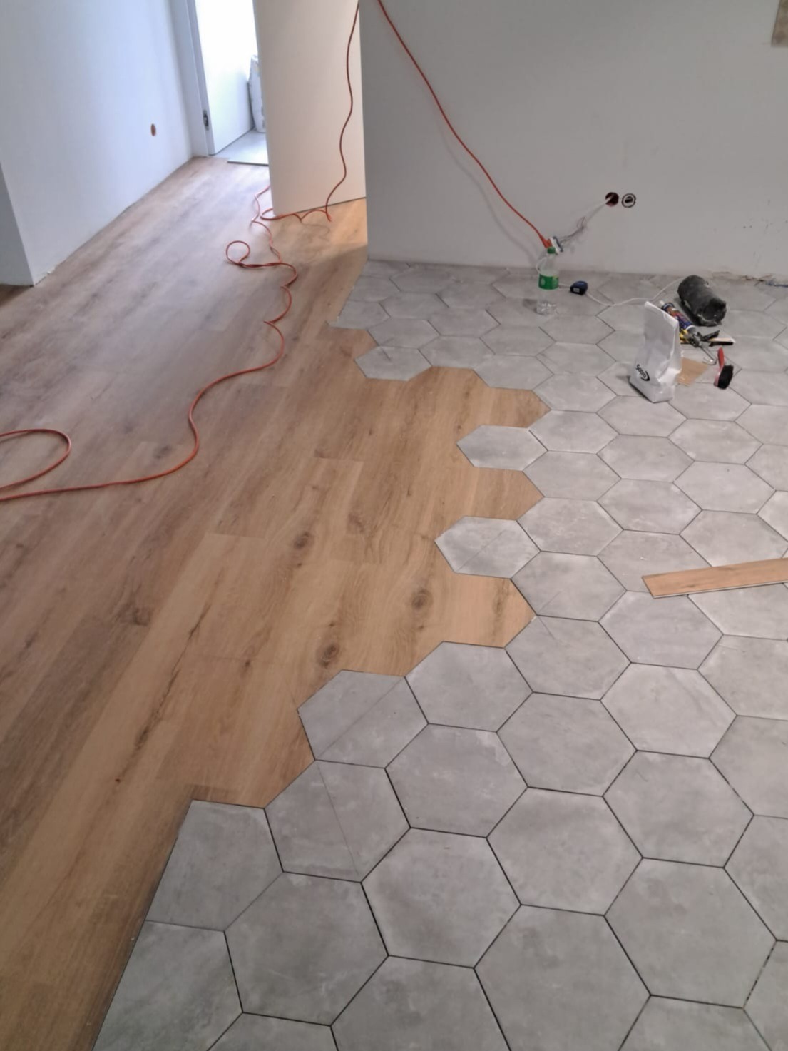 Układanie szarych, heksagonalnych płytek na podłodze, częściowo położone obok paneli podłogowych imitujących drewno; widoczne narzędzia glazurnicze i kabel zasilający w tle.