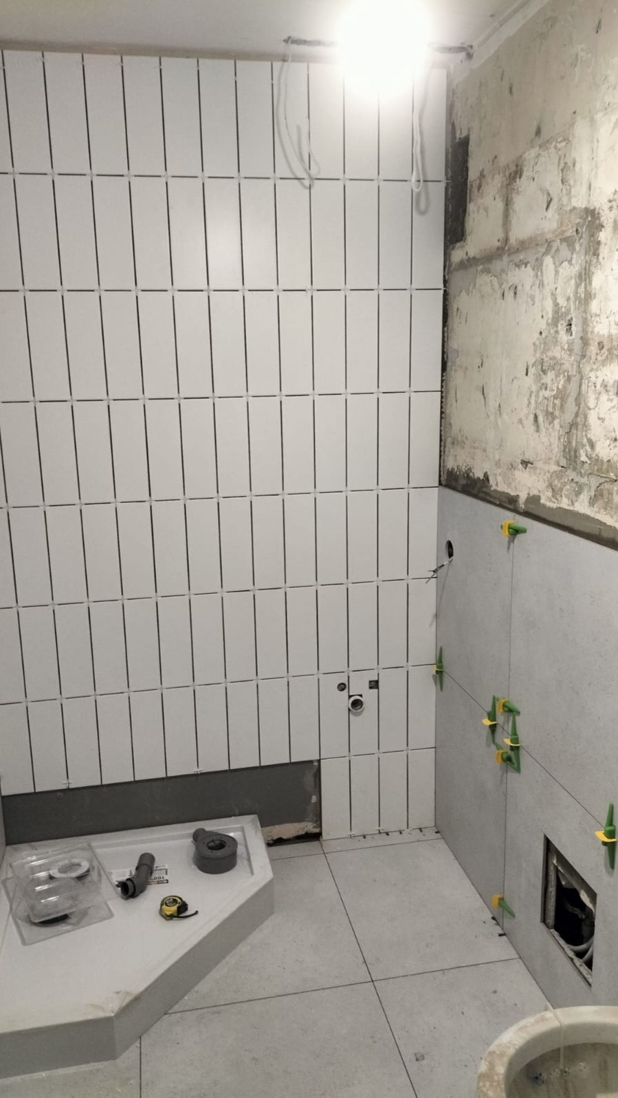 Łazienka w trakcie remontu: białe, pionowe płytki na ścianie z czarną fugą, szare płytki na podłodze i ścianie, widoczne akcesoria instalacyjne oraz poziomica, fragment ściany z surowego betonu.