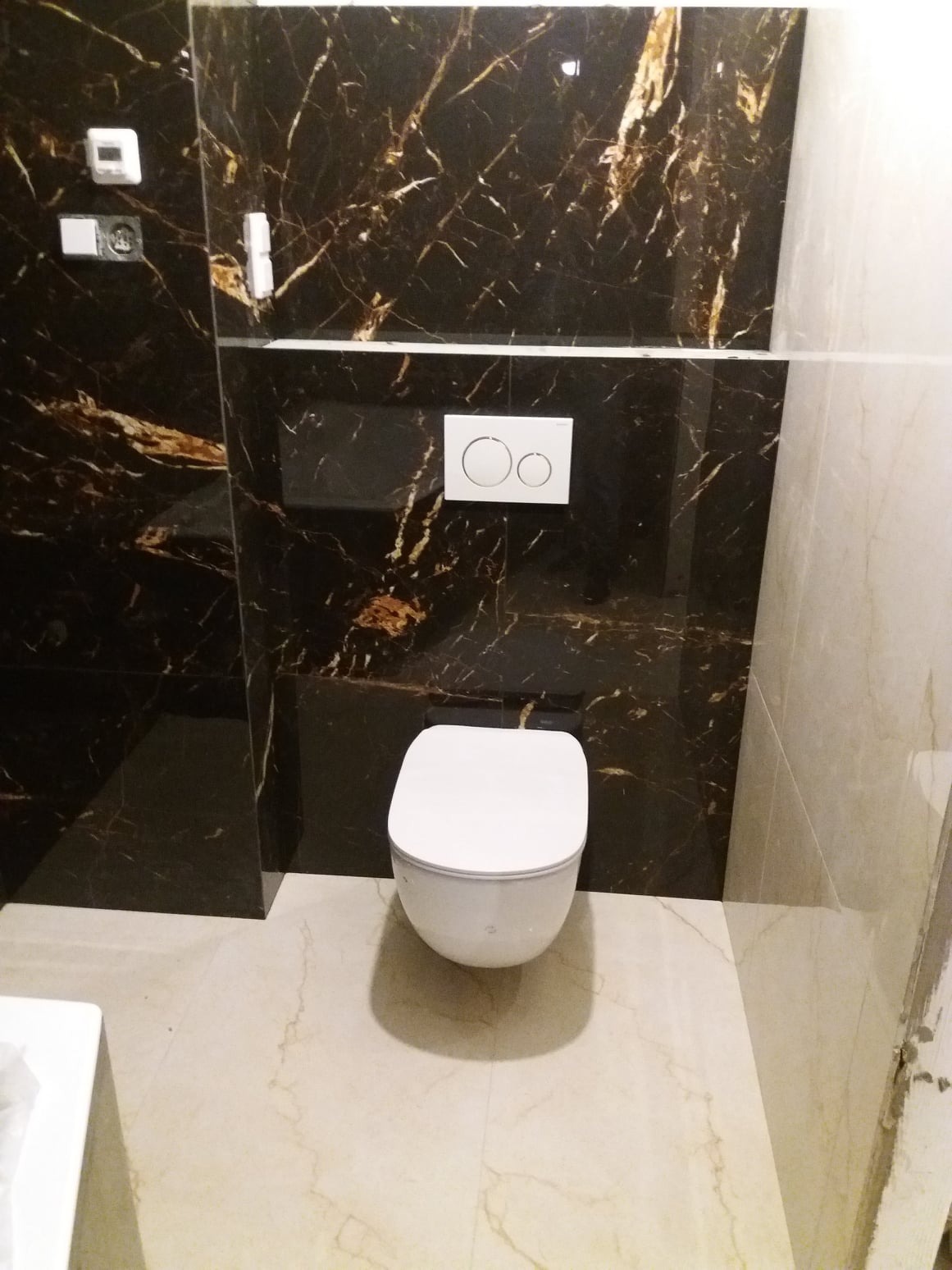Nowoczesna toaleta podwieszana na tle ściany z czarnego marmuru z białymi żyłami, biały przycisk spłuczki, jasna marmurowa podłoga.