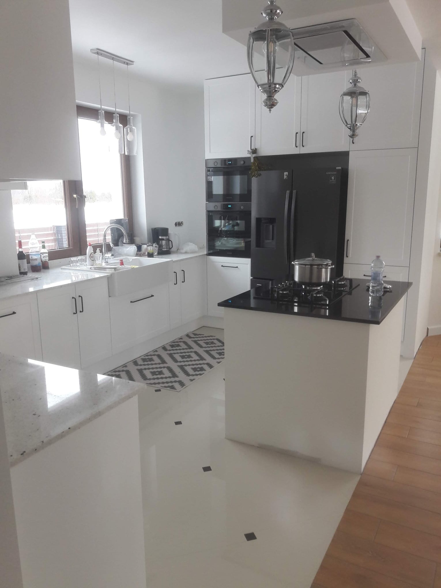 Nowoczesna, jasna kuchnia z białymi szafkami, czarnym sprzętem AGD i wyspą kuchenną z czarnym blatem, oświetlona lampami wiszącymi w stylu retro.