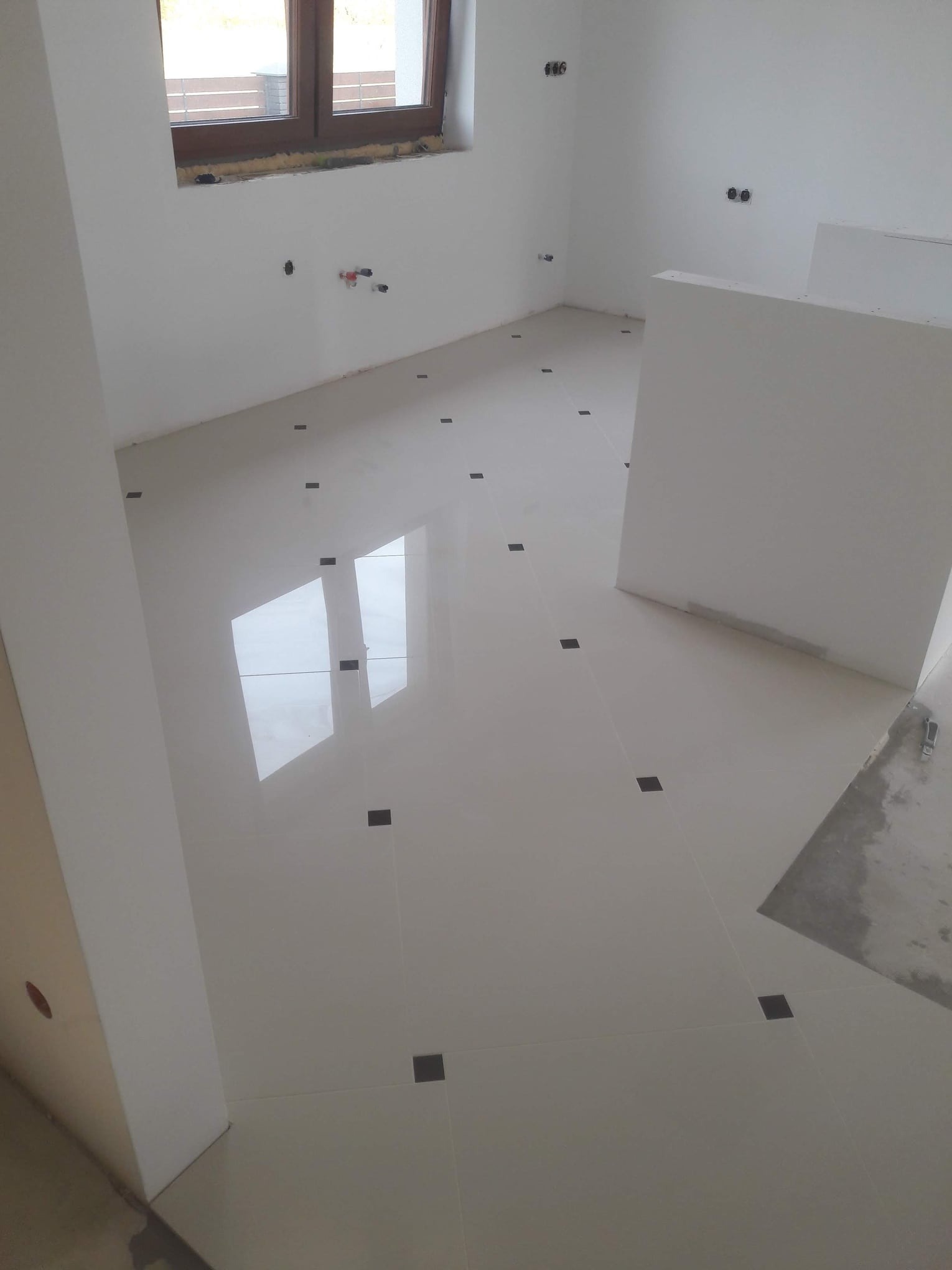 Błyszcząca, kremowa podłoga z płytek z czarnymi akcentami w nowym wnętrzu. Widoczne przygotowane punkty instalacji na ścianach i okno z pianką montażową.