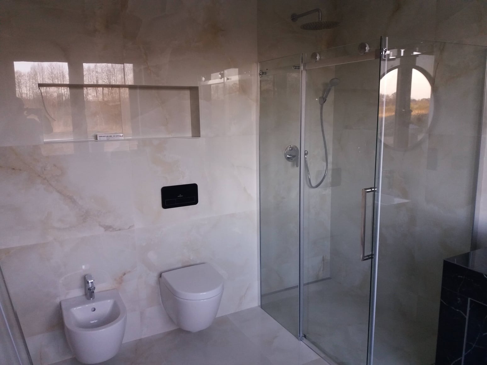 Nowoczesna łazienka z beżowymi marmurkowymi płytkami, wisząca toaleta i bidet, kabina prysznicowa ze szklanymi drzwiami przesuwnymi i okrągłym oknem odbijającym krajobraz na zewnątrz.