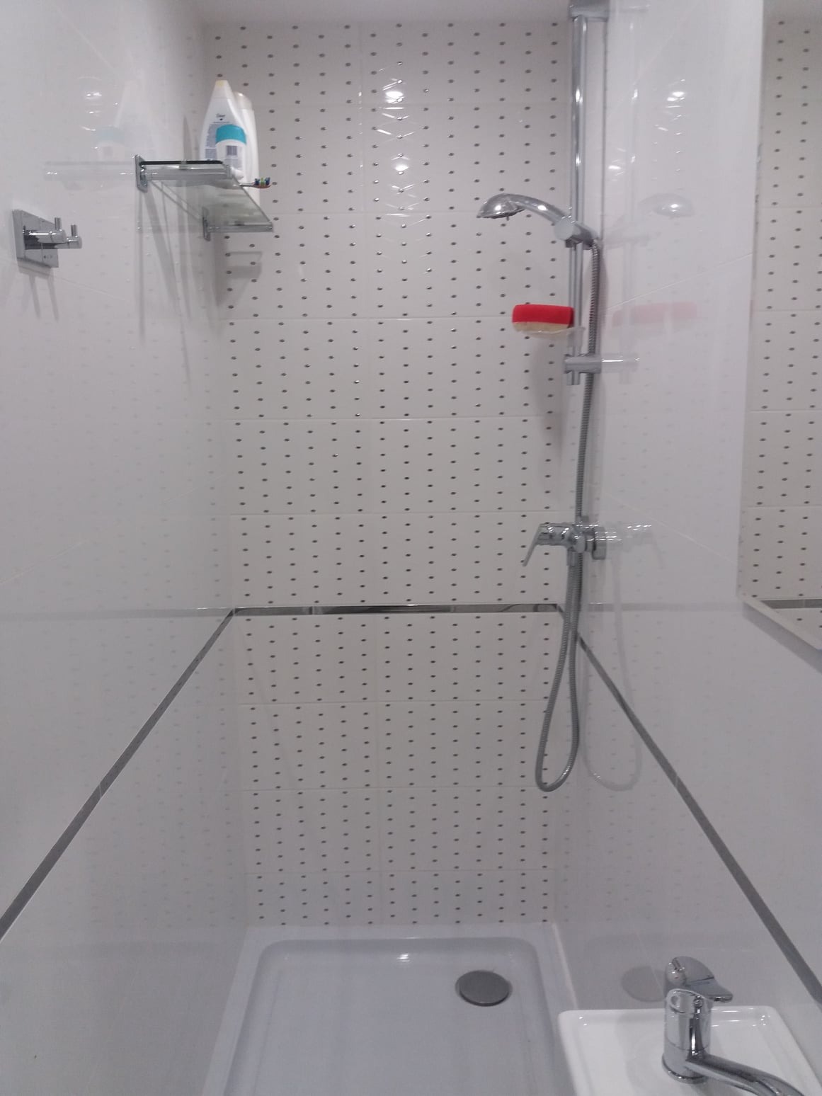 Nowoczesna kabina prysznicowa z białymi płytkami w kropki, chromowaną armaturą i szklaną półką z kosmetykami.