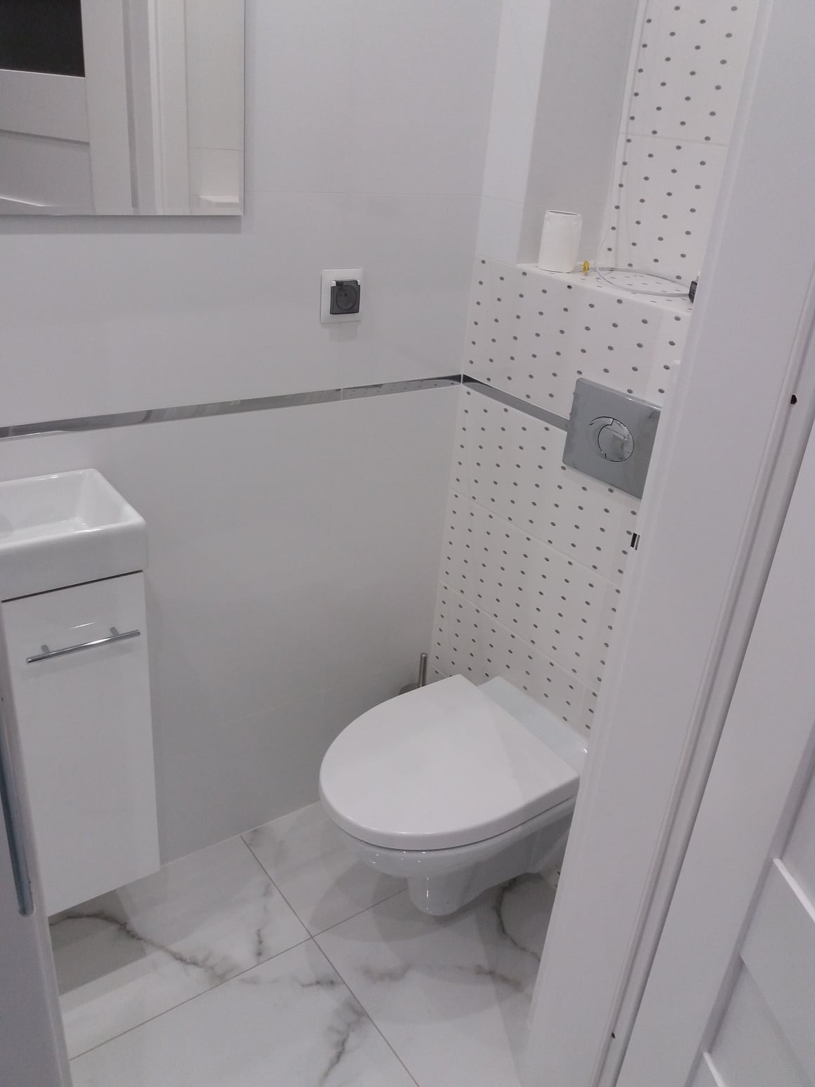 Nowoczesna, jasna toaleta z podtynkową spłuczką, umywalką z szafką i płytkami w kropki na ścianie, widoczna z narożnika pomieszczenia.