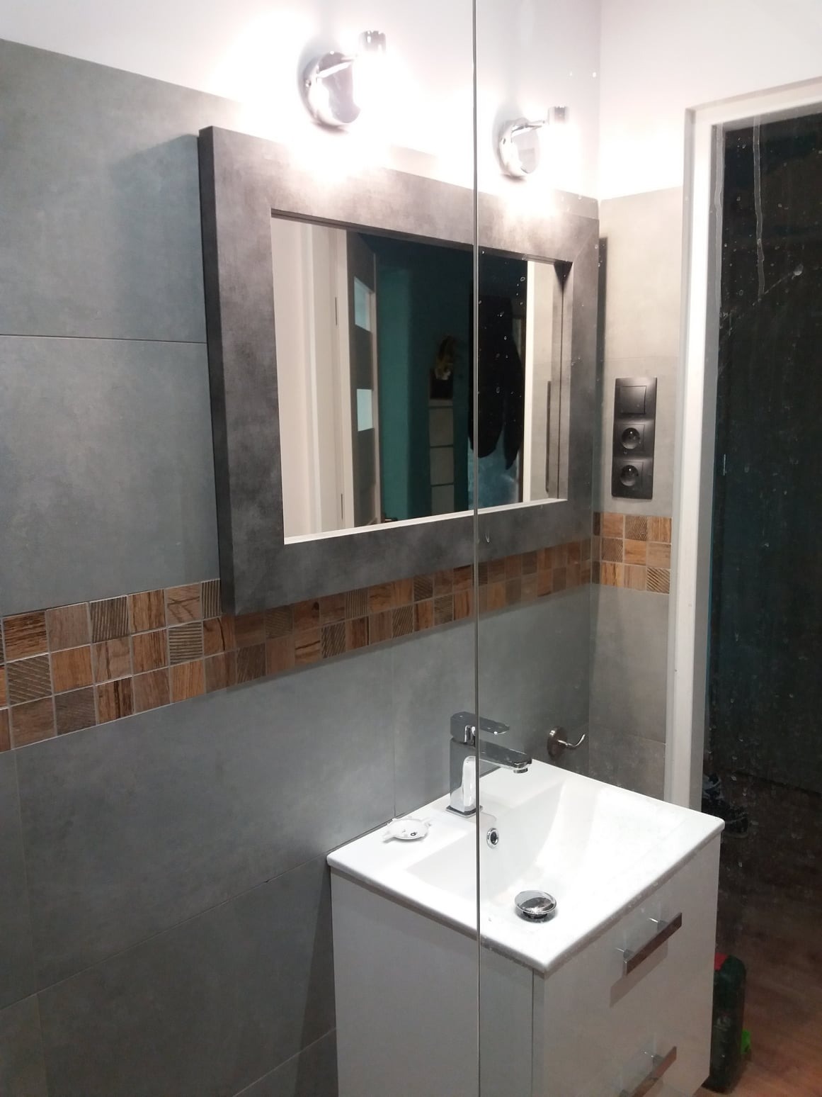 Nowoczesna łazienka z szarymi płytkami, dekoracyjnym pasem brązowych mozaiek, umywalką z szafką i lustrem w szarej ramie oświetlonym dwoma lampami.