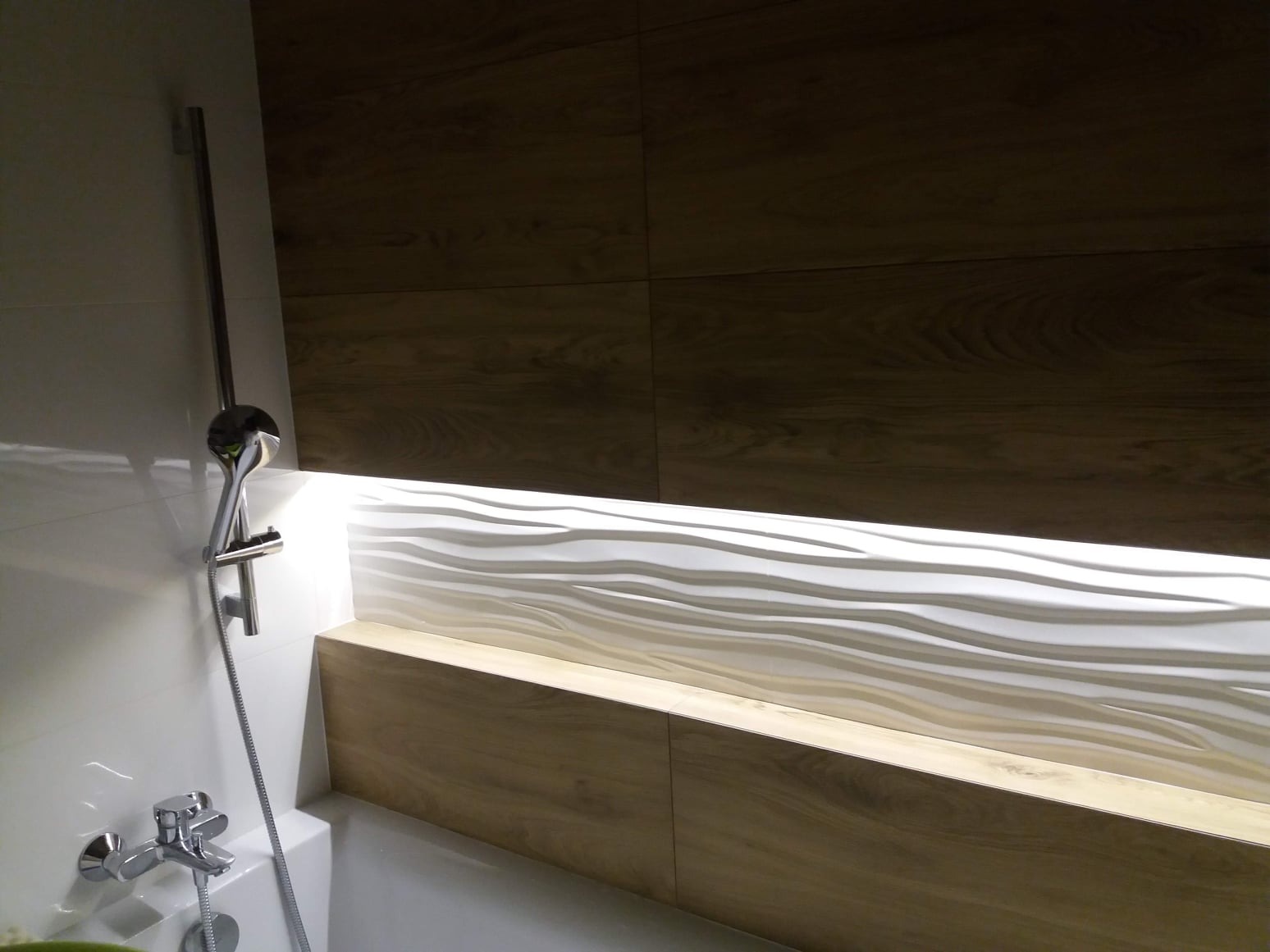 Nowoczesna łazienka z drewnopodobnymi płytkami, dekoracyjną białą falistą ścianą z podświetleniem LED oraz chromowaną baterią i słuchawką prysznicową.