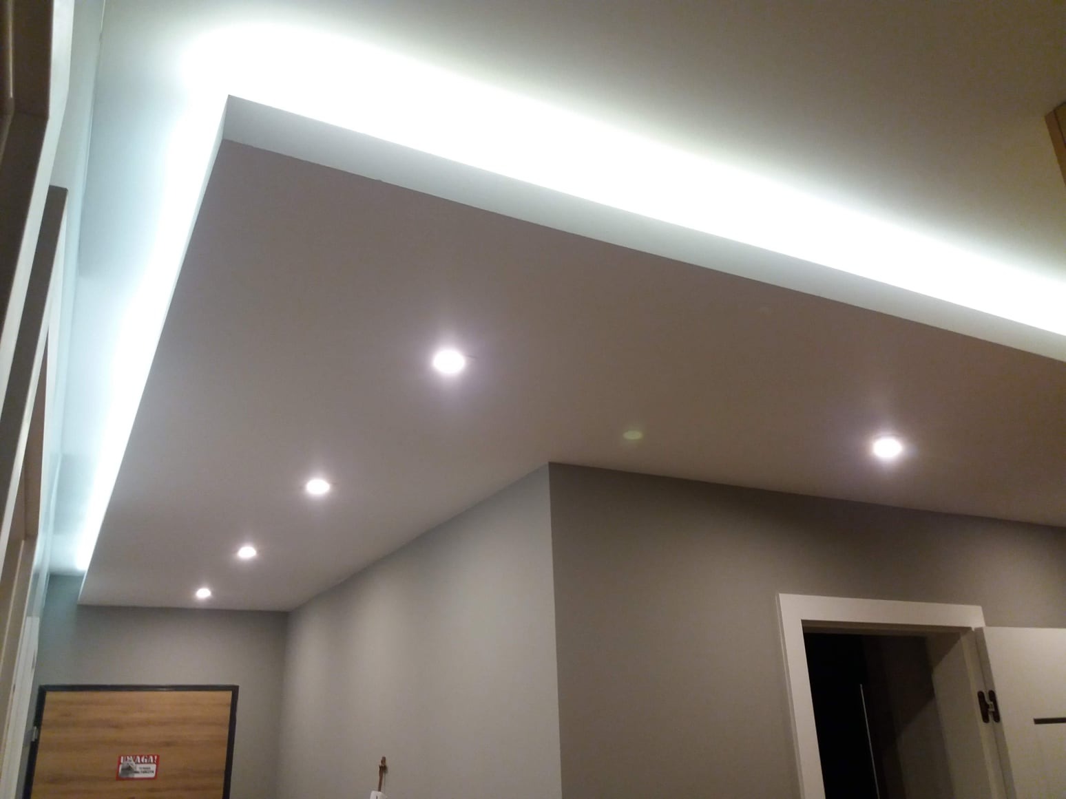 Sufit podwieszany z oświetleniem LED: listwa LED wzdłuż krawędzi i rząd punktowych lamp halogenowych, szara ściana z drzwiami w tle.