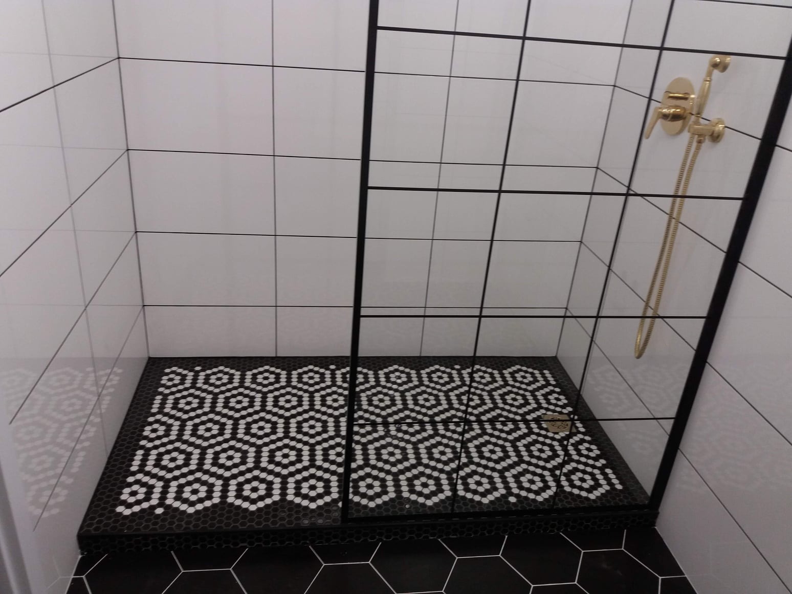 Nowoczesna kabina prysznicowa z białymi kwadratowymi płytkami na ścianach i podłogą wyłożoną czarno-białą mozaiką w kształcie heksagonów, złota bateria prysznicowa.