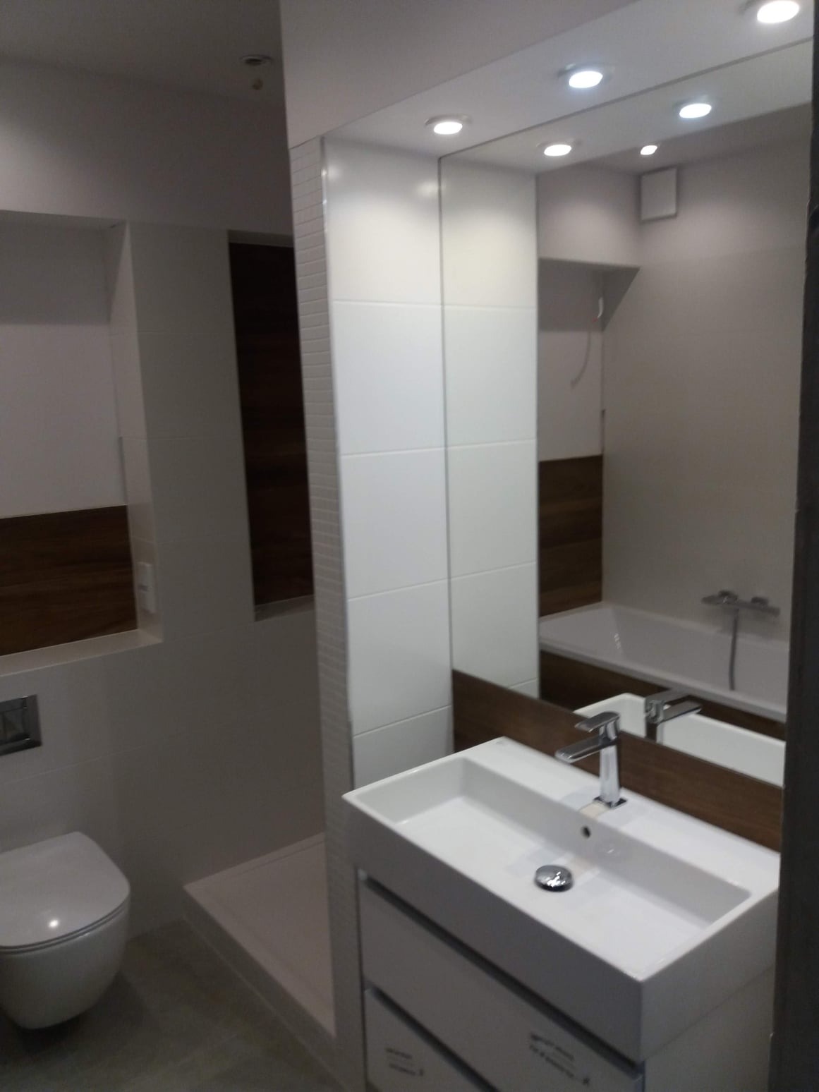 Nowoczesna łazienka z białą umywalką nablatową na szafce, duże lustro z oświetleniem punktowym, widoczna toaleta i fragment kabiny prysznicowej, ściany wykończone białymi płytkami i panelami...