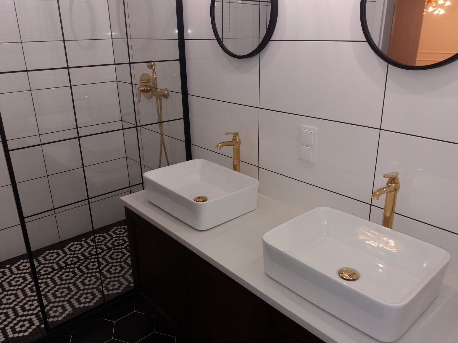 Nowoczesna łazienka z dwoma białymi umywalkami nablatowymi, złotymi bateriami i czarnymi okrągłymi lustrami na ścianie z białych płytek z czarną fugą. W tle widoczny prysznic ze złotą armaturą...