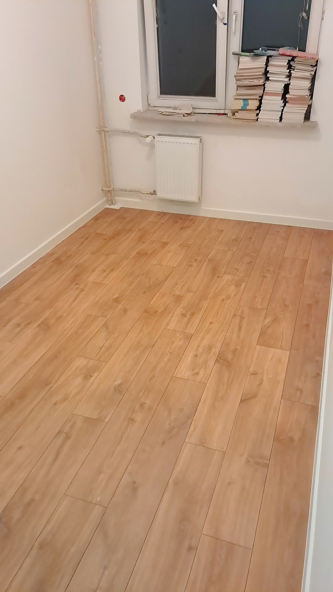 Nowo położona podłoga panelowa w jasnym odcieniu drewna, widoczny grzejnik pod oknem i stos książek na parapecie, ściany pomalowane na biało.