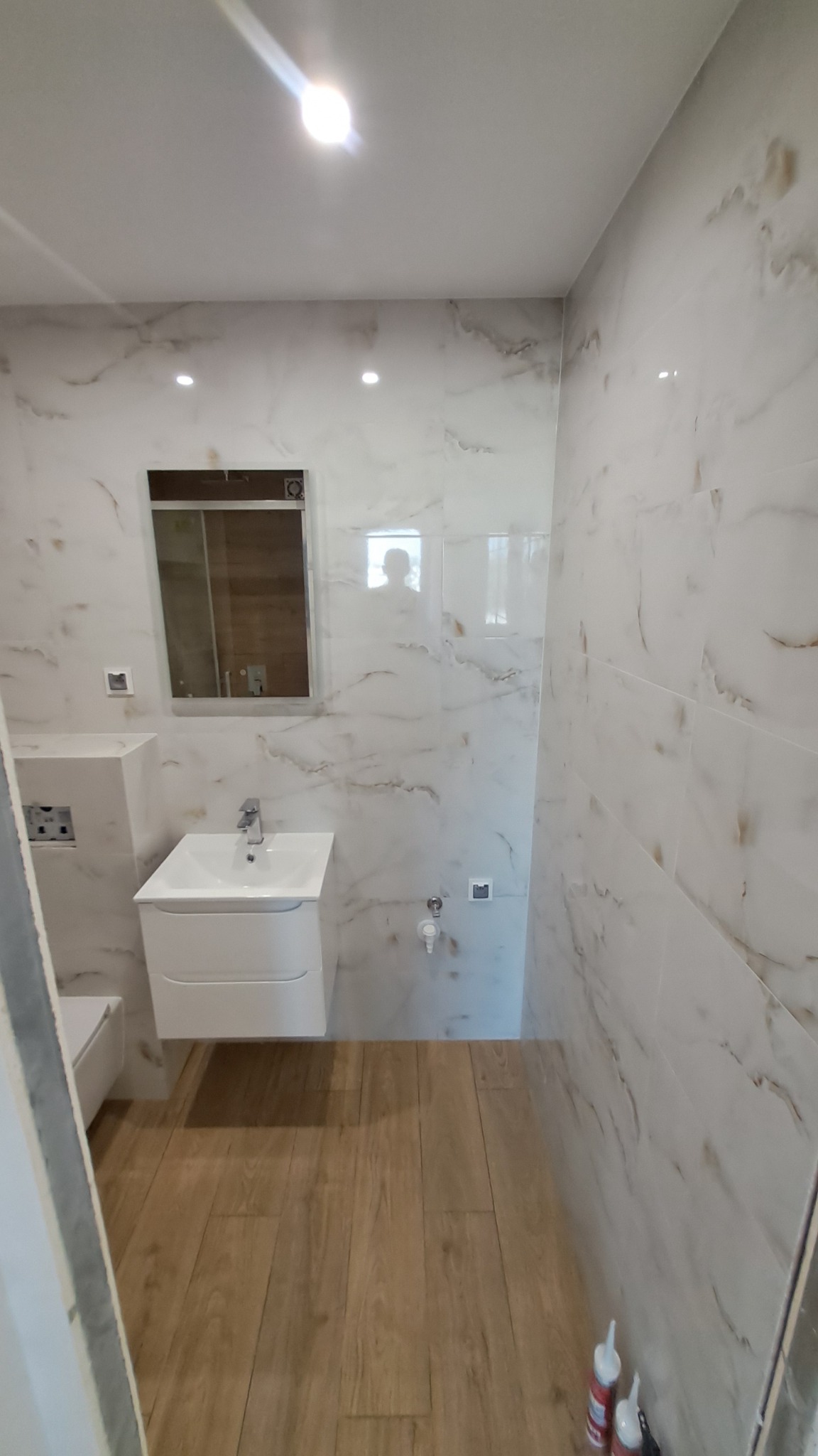 Nowoczesna łazienka z białymi płytkami imitującymi marmur i drewnianą podłogą, umywalka z szafką w minimalistycznym stylu, widoczne silikony budowlane w rogu pomieszczenia.