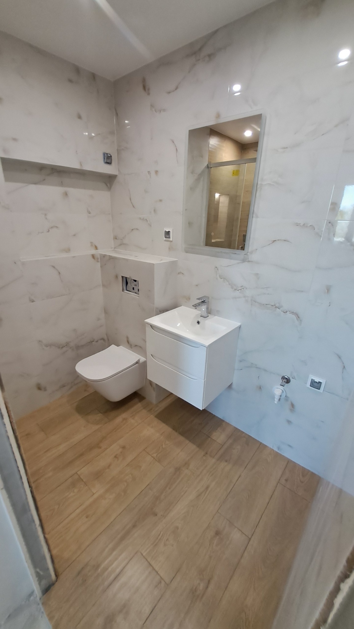 Nowoczesna łazienka z białymi płytkami imitującymi marmur, drewnopodobną podłogą, podwieszaną toaletą i szafką z umywalką, widok na kabinę prysznicową w lustrze.