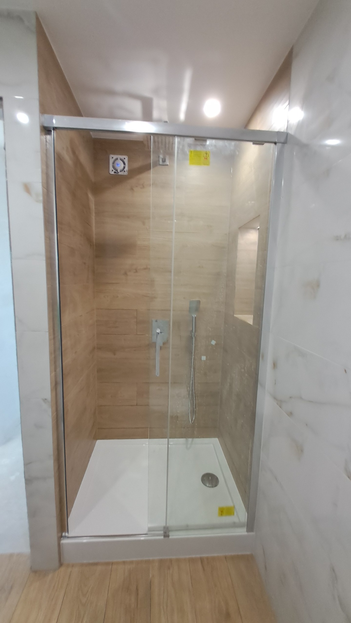 Kabina prysznicowa z przesuwnymi drzwiami, wykończona płytkami imitującymi drewno na ścianach i marmurem na sąsiedniej ścianie, widoczny odpływ w brodziku i panel wentylacyjny w górnej części kabiny.