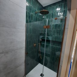 Paweł Tumel - Nowoczesna kabina prysznicowa z zielonymi, pionowymi płytkami i szklaną ścianką, wykończona elementami w kolorze miedzianym. Szare płytki na sąsiedniej ścianie.