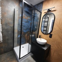 Paweł Tumel - Nowoczesna łazienka z kabiną prysznicową typu walk-in z czarnymi ramami, umywalką nablatową i lustrem w owalnej ramie na tle dekoracyjnej ściany.