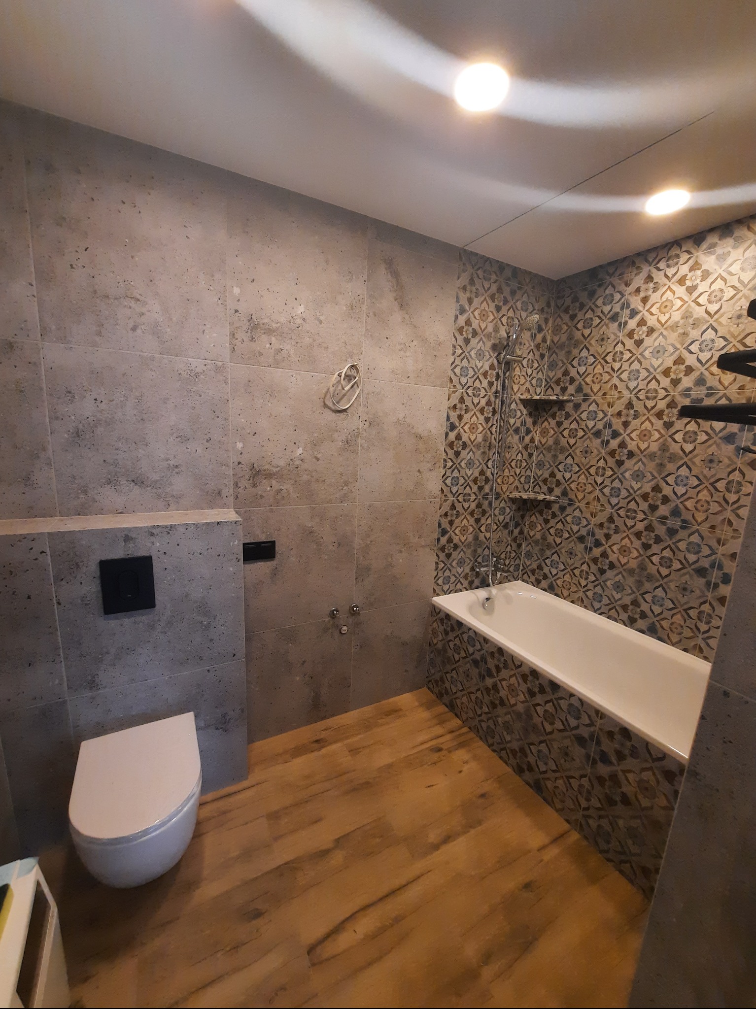 Wykończona łazienka z drewnopodobną podłogą i mozaiką przy wannie. Ściany wyłożone płytkami imitującymi kamień. Wisząca toaleta i minimalistyczny design.