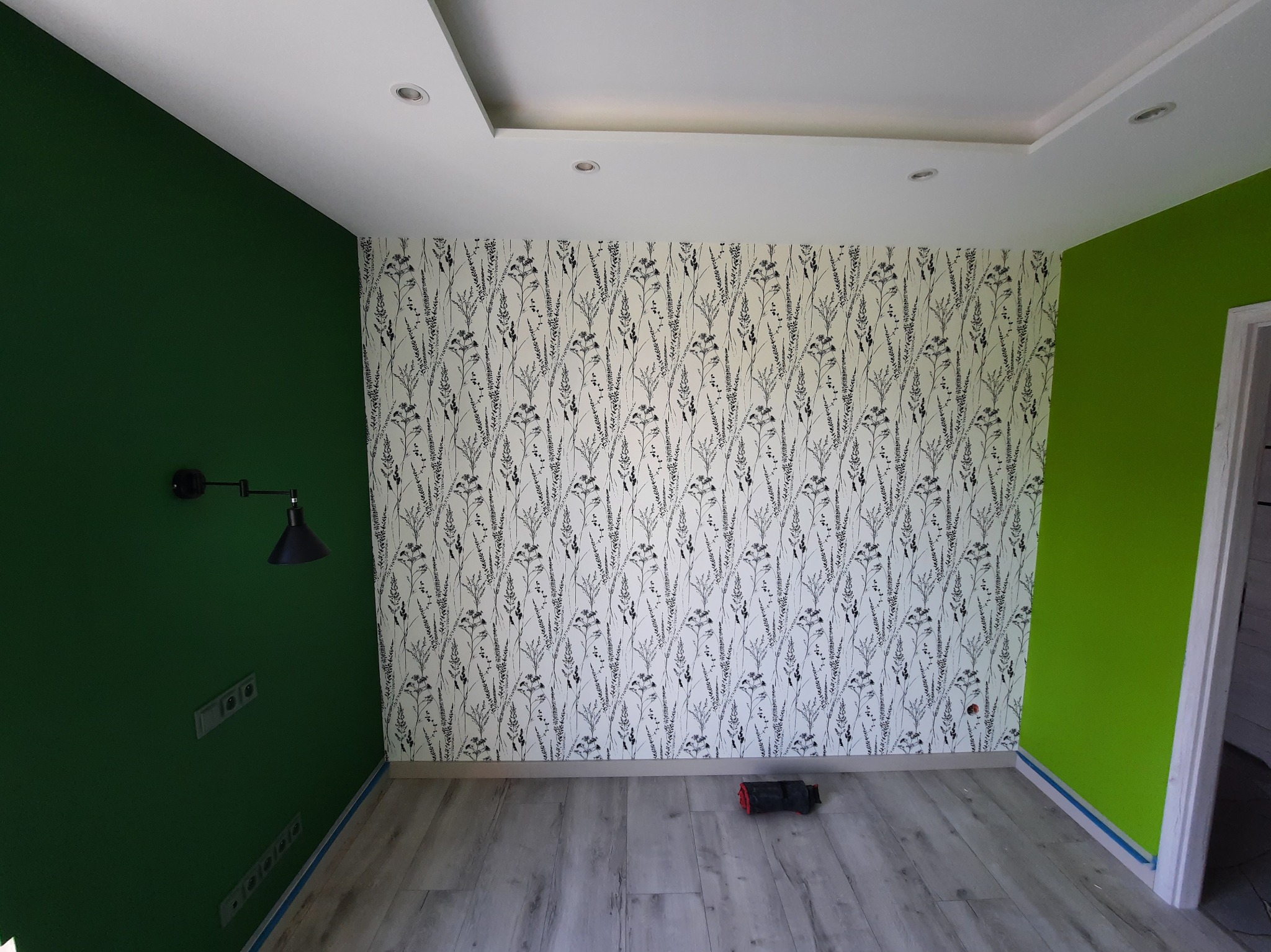 Wnętrze pokoju po remoncie: ściana w zielonym kolorze, ściana w białe wzory roślinne, szara podłoga. Widoczna lampa i gniazdka elektryczne.