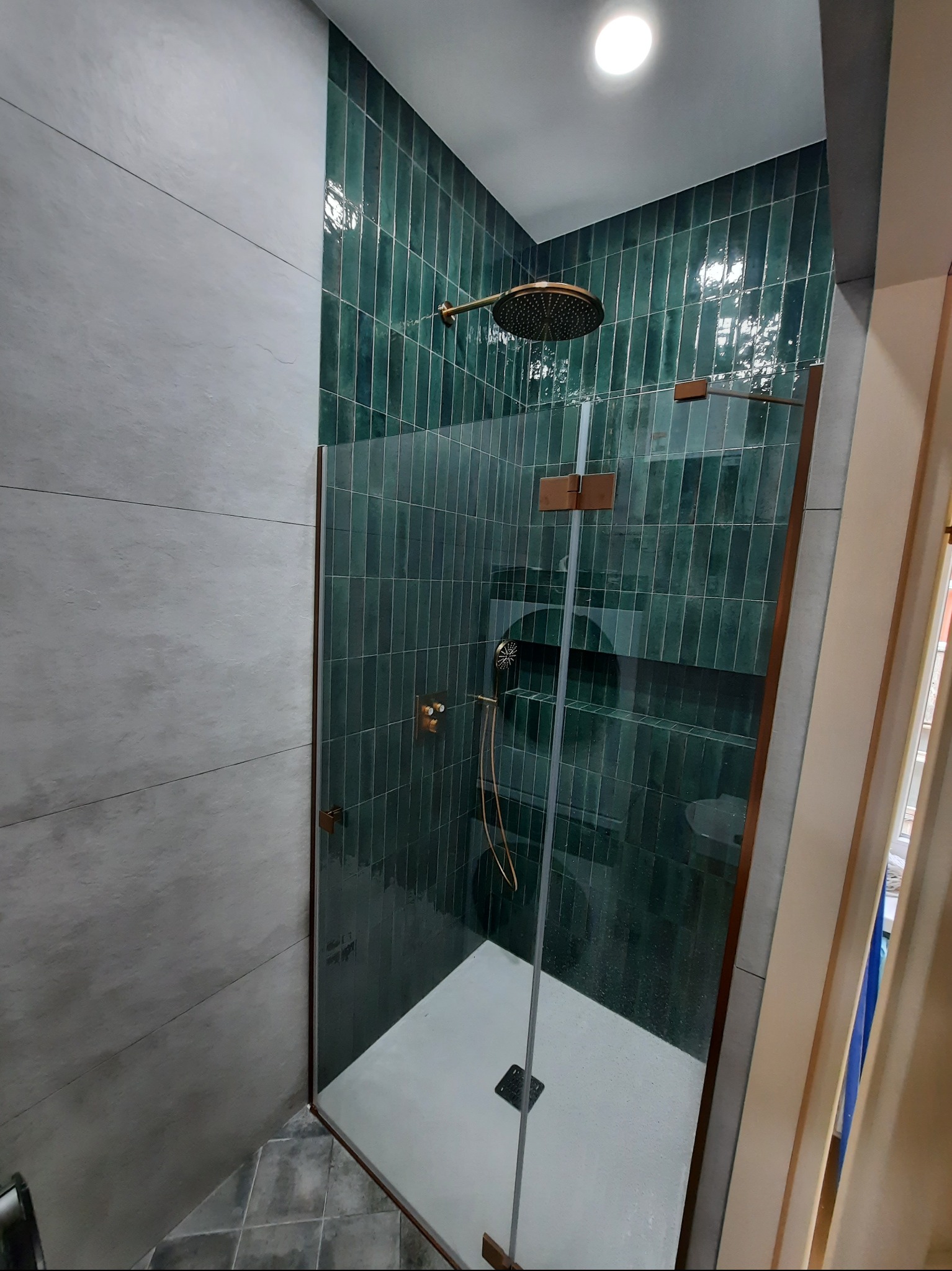 Nowoczesna kabina prysznicowa z zielonymi, pionowymi płytkami i szklaną ścianką, wykończona elementami w kolorze miedzianym. Szare płytki na sąsiedniej ścianie.