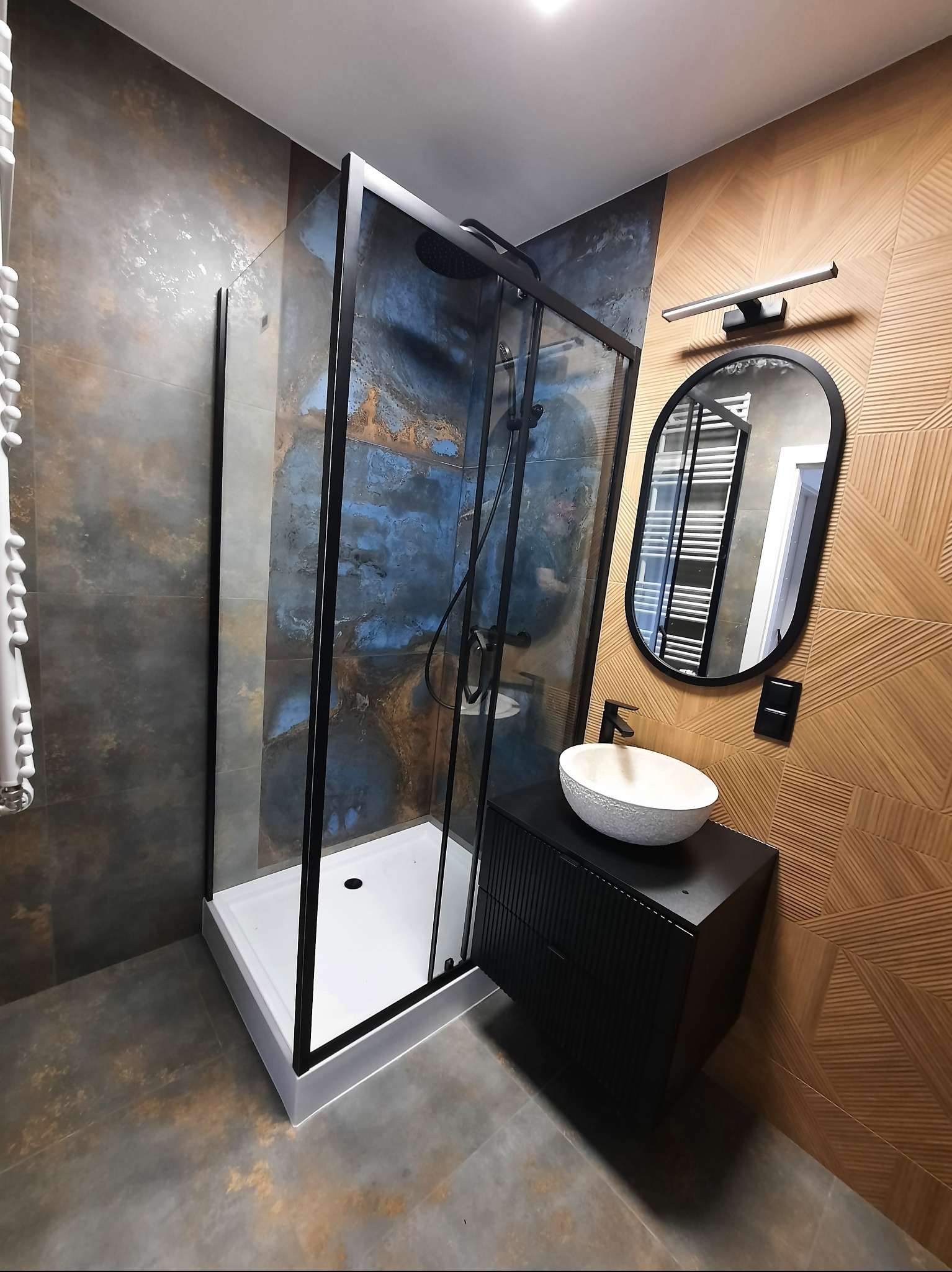 Nowoczesna łazienka z kabiną prysznicową typu walk-in z czarnymi ramami, umywalką nablatową i lustrem w owalnej ramie na tle dekoracyjnej ściany.