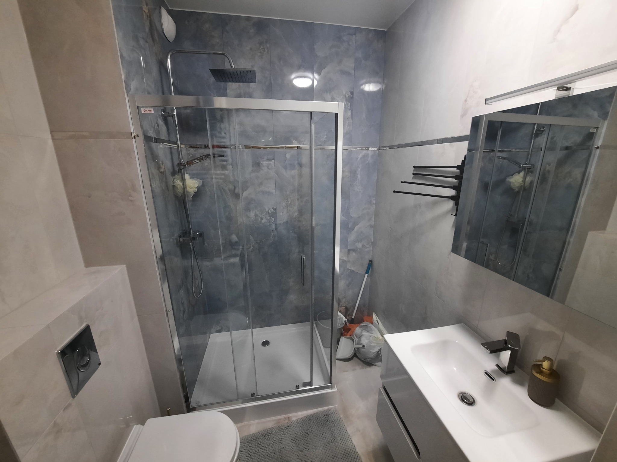 Wykończona łazienka z kabiną prysznicową typu walk-in, umywalką z szafką i lustrem. Płytki imitujące marmur na ścianach. Widoczne akcesoria łazienkowe.