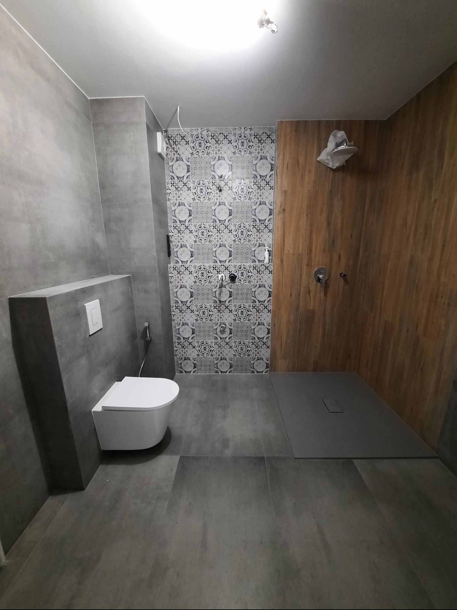 Nowoczesna łazienka z szarymi płytkami, kabiną prysznicową wykończoną drewnem i wzorzystymi płytkami. Wisząca toaleta. Minimalistyczny design.