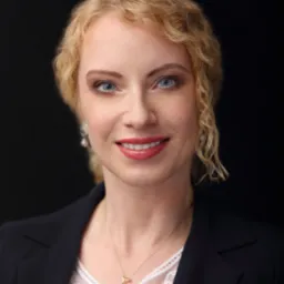 Portret uśmiechniętej kobiety w czarnej marynarce na ciemnym tle. Profesjonalny wizerunek adwokata z Warszawy, z blond włosami i delikatną biżuterią.